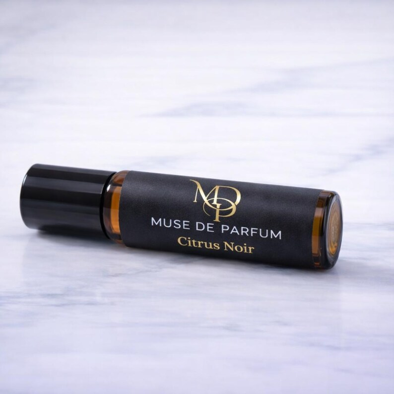 K&ouml;nnte beinhalten: Eine bernsteinfarbene Roll-on-Parf&uuml;mflasche mit schwarzem Verschluss und dunklem Etikett. Das Etikett tr&auml;gt den goldenen Text "MUSE DE PARFUM Citrus Noir". Die Flasche liegt auf einer wei&szlig;en Marmoroberfl&auml;che.