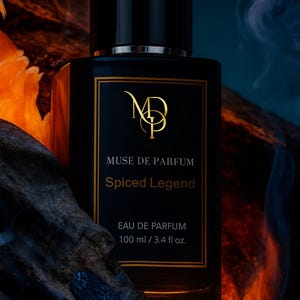 May include: A black rectangular perfume bottle with a gold label. The label reads "MUSE DE PARFUM Spiced Legend EAU DE PARFUM 100 ml / 3.4 fl oz." The bottle has a black cap and sits on a dark, textured surface with an orange and blue background.