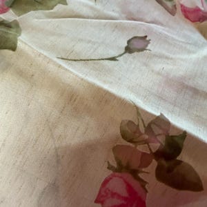 May include: Close-up of a fabric with a floral pattern. The fabric is a light beige color, with pink roses and green leaves. The roses are in various stages of bloom, and the leaves have a watercolor effect.