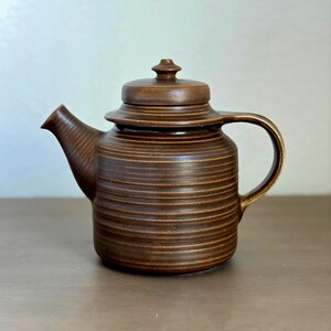 May include: A dark brown ceramic teapot featuring a cylindrical body with horizontal ribbed textures, a matching lid with a small knob handle, a curved spout, and a sturdy side handle. The teapot has a uniform matte finish throughout its exterior.