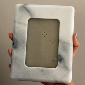 May include: A rectangular picture frame crafted from white marble with prominent grey veining throughout. The frame features a smooth, polished finish and a central rectangular opening designed to hold a photograph or display item.