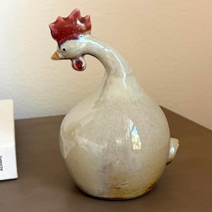 May include: A ceramic chicken figurine with a glossy, off-white, rounded body and a long, curved neck. The head features a bright red comb, a small orange beak, and a red wattle. The base of the figurine shows subtle brown and tan glaze variations.