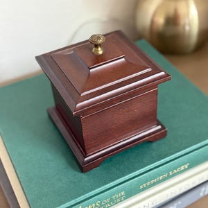 May include: A small, square, dark brown wooden box with a decorative brass knob on top. The box is sitting on top of a stack of books with green and gray covers. The box has a classic, vintage aesthetic.