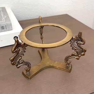 May include: A brass display stand featuring a circular top ring supported by three legs shaped like stylized dragons. The metal has a weathered, antique finish with visible texture and patina throughout the structure, providing a stable base.