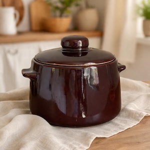 May include: A dark brown ceramic cooking pot with a lid, set on a wooden surface covered with a beige cloth. The pot has two side handles and a rounded lid. The kitchen background includes wooden cutting boards and plants.