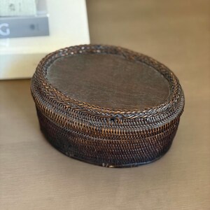 May include: A woven brown oval basket with a dark wooden top. The basket has a braided rim and a textured, woven body. The basket is sitting on a light brown surface. The basket is likely used for storage or decoration.