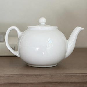 May include: A glossy white ceramic teapot with a rounded body, a curved handle on the left side, a matching lid with a small knob handle on top, and a tapered spout on the right side. The teapot has a smooth, solid finish and a classic shape.