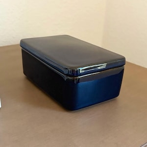 May include: A rectangular, deep navy blue ceramic butter dish with a smooth, glossy finish. The container features a matching lid with slightly rounded edges and a clean, minimalist design, showcasing a solid, dark color throughout the piece.