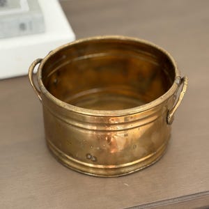 May include: A round brass basin with a polished metallic finish, featuring two small curved handles on opposite sides and horizontal ribbed detailing around the exterior wall of the vessel, showing a warm golden tone and a smooth texture.