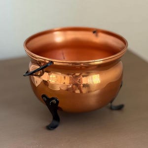 May include: A polished copper cauldron with a rounded body and a wide, open top. The vessel features three curved black metal legs attached to the base and a black metal handle attached to the sides, providing a sturdy structure for this container.