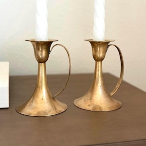May include: Two brass candle holders with flared bases and curved side handles. Each holder features a tall, slender stem and a wide cup at the top, currently holding a white, spiraled, tapered candle. The brass has a warm, metallic finish.
