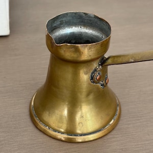 May include: A brass coffee pot with a wide base, a narrow neck, and a flared rim. The interior is lined with a silver-colored metal. A long, flat handle is attached to the side with two visible copper rivets. The exterior surface shows signs of wear.