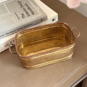 May include: A rectangular brass planter with a ribbed design and small handles. The planter sits on a wooden surface next to a stack of books titled "The Interior Design Handbook."
