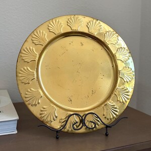 May include: A round, metallic gold-toned decorative plate featuring a raised, embossed shell pattern along the wide rim. The center of the plate has a smooth, reflective surface with visible signs of wear and minor surface scratches throughout.