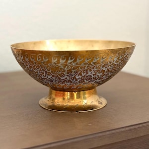 May include: A brass bowl with a wide, open top and a pedestal base. The exterior surface features an intricate, etched floral pattern with dark, recessed lines contrasting against the polished gold-toned metal of the bowl and the base.