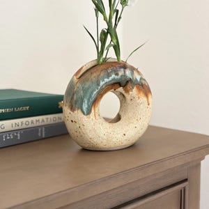 May include: A speckled ceramic vase with a unique donut shape, featuring a hole in the center. The vase has a brown, green, and blue glaze and holds white flowers with green stems. The vase is on a wooden surface.