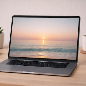Puede incluir: Un portátil plateado que muestra una escena de puesta de sol en la playa. Una taza blanca, un ratón y una pequeña planta en maceta están sobre un escritorio de madera. También hay una pila de cuadernos grises en el escritorio.