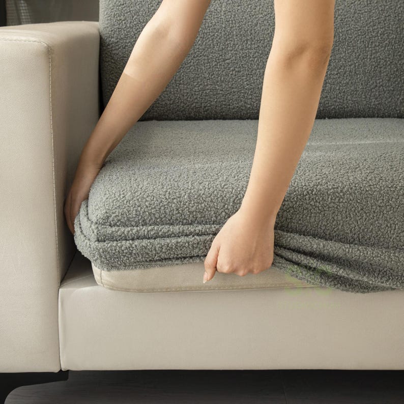May include: A gray, textured, fleece-like sofa cover featuring a thick, curly pile fabric. The cover is fitted over a light-colored, smooth-textured sofa cushion, with the fabric edges visible along the sides of the seat and backrest.