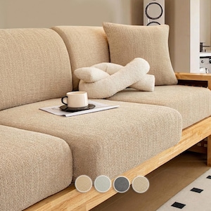 Pode incluir: Um sofá seccional bege com estrutura de madeira. Possui tecido texturizado, uma almofada quadrada e uma almofada decorativa em forma de nó. Uma xícara de café está em uma revista no sofá. O sofá é de cor neutra.