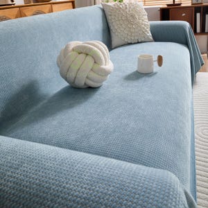 Puede incluir: Un sofá azul claro con una manta a juego. Un cojín decorativo blanco con forma de nudo y una taza blanca con asa de madera están en el sofá. También hay un cojín floral decorativo en el sofá.