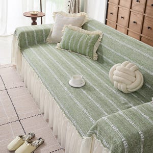 May include: A green sofa cover with a white striped pattern and a white ruffled skirt. The sofa is adorned with decorative pillows in cream and green, and a cream-colored knot-shaped pillow. A teacup and slippers are also visible.