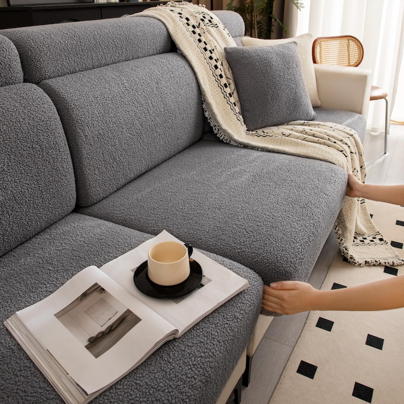 May include: A gray textured sofa cover made of a soft, fuzzy fabric. The cover features a thick, plush pile and is draped with a cream-colored throw blanket with black geometric patterns. A matching gray square pillow rests on the sofa seat.