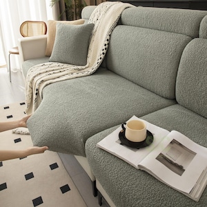 May include: A sage green textured sherpa sofa cover featuring a thick, plush fabric. The cover is draped over a sectional sofa, accompanied by a matching square throw pillow and a cream-colored knit throw blanket with a black geometric pattern.