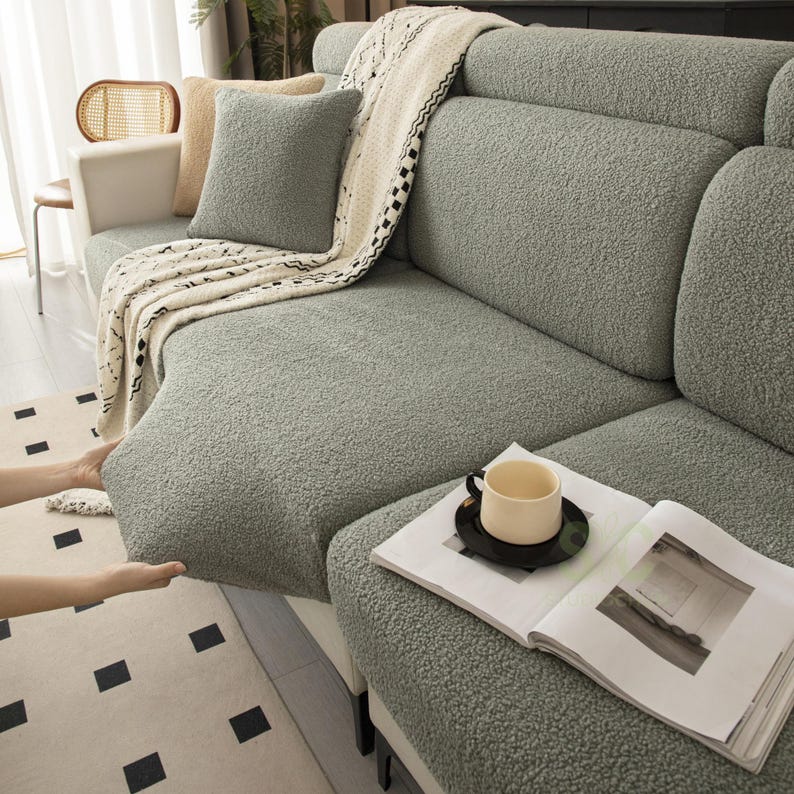 May include: A sage green textured sherpa sofa cover featuring a thick, plush fabric. The cover is draped over a sectional sofa, accompanied by a matching square throw pillow and a cream-colored knit throw blanket with a black geometric pattern.