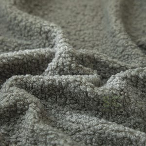 May include: A grey fabric with a thick, curly, and textured pile surface. The material features a dense, looped construction that creates a soft, uneven appearance with deep folds and shadows across the surface of the textile.