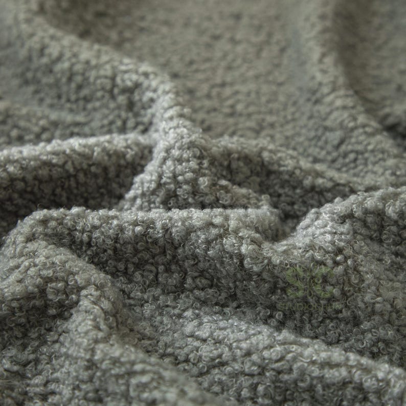 May include: A grey fabric with a thick, curly, and textured pile surface. The material features a dense, looped construction that creates a soft, uneven appearance with deep folds and shadows across the surface of the textile.