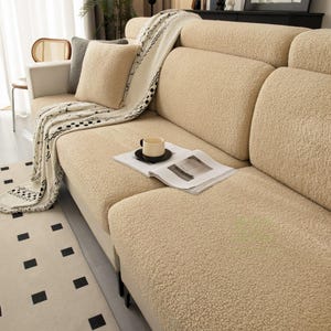 May include: A beige textured sherpa sofa cover featuring a thick, plush fabric. The set includes a matching beige throw pillow and a cream-colored throw blanket with a black and white geometric pattern, draped over the seat cushions.