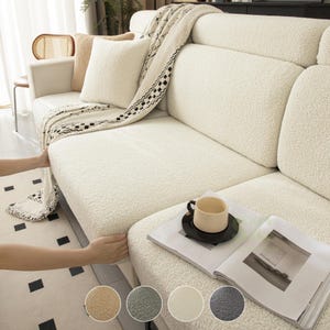 May include: A cream-colored, textured sherpa sofa cover draped over a couch. The cover features a soft, fleece-like material with a nubby surface. A matching throw blanket with a black and white geometric pattern is layered on top of the sofa.