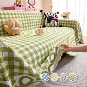 Puede incluir: Una funda de sofá a cuadros verdes y blancos con un borde con flecos. La funda está colocada sobre un sofá, con un cojín decorativo y una bola de peluche visibles. La funda está hecha de una tela suave y tejida.
