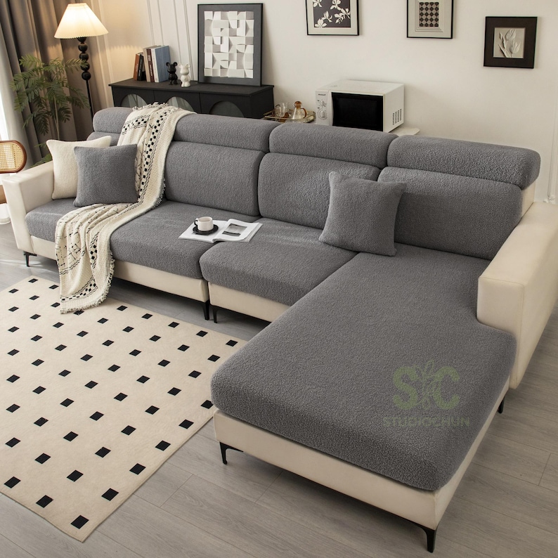 May include: A sectional sofa featuring a textured, dark gray fabric cover on the seat cushions and backrests, contrasted by a light cream-colored base and armrests. The sofa includes two matching gray throw pillows and a white patterned throw blanket.