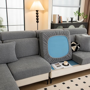 Pode incluir: Um sofá seccional cinza com uma capa de almofada azul clara parcialmente removida, revelando um interior azul. Uma almofada decorativa em preto e branco e uma xícara de café pequena estão em uma mesa branca. O sofá tem um design moderno.