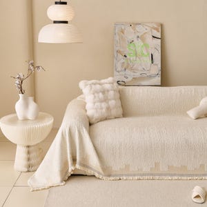 Könnte beinhalten: Ein cremefarbenes Sofa mit einer strukturierten Tagesdecke und dekorativen Kissen. Ein weißes, flauschiges Kissen liegt auf dem Sofa. Ein modernes, abstraktes Gemälde hängt an der Wand. Eine weiße Papierlampe und ein kleiner Beistelltisch mit einer Vase befinden sich ebenfalls im Raum.