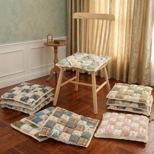 Puede incluir: Una silla de madera con un cojín cuadrado acolchado y varios otros cojines cuadrados en varios colores y patrones. Los cojines están hechos de tela de retazos y tienen un ribete de encaje. La silla y los cojines están sobre un piso de madera noble.