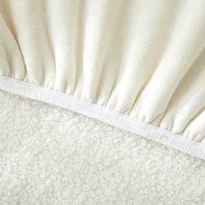 May include: A cream-colored fabric with a textured, woven surface featuring gathered folds along a white elastic band. The material transitions into a dense, curly, off-white fleece-like texture below the elastic edge, creating a contrast.