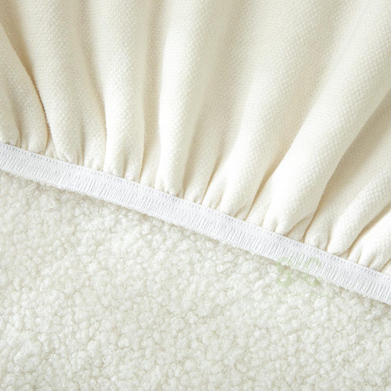 May include: A cream-colored fabric with a textured, woven surface featuring gathered folds along a white elastic band. The material transitions into a dense, curly, off-white fleece-like texture below the elastic edge, creating a contrast.