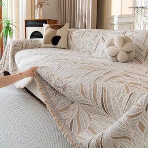 Peut inclure: Housse de canapé beige à motif de feuilles. La housse est drapée sur un canapé blanc, avec une frange sur le bord. Des coussins décoratifs dans les tons beige et marron ajoutent à l'esthétique cosy. Une main est montrée en train d'ajuster la housse.