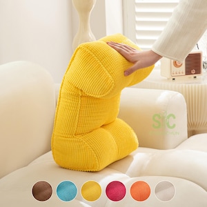 Può includere: Un cuscino da lettura in velluto a coste giallo brillante con un design strutturato a più sezioni. Il tessuto presenta una distinta trama a coste verticali. Il cuscino è posizionato su un divano di colore chiaro, mostrandone la forma ergonomica.