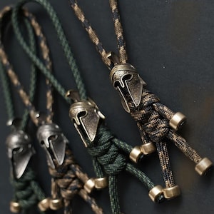May include: Close-up of several Spartan helmet keychains. Each keychain features a bronze-colored helmet pendant attached to a braided cord in various camouflage patterns, including green, brown, and black. The keychains have bronze-colored metal accents.