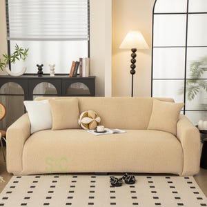 Op de afbeelding: Een beige, getextureerde bankhoes van zacht, fleece-achtig materiaal. De hoes is over een driezitsbank geplaatst en bevat twee bijpassende beige sierkussens, &eacute;&eacute;n wit sierkussen en een klein, rond decoratief kussen.