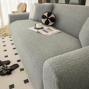 Op de afbeelding: Een lichtgrijze bankhoes met een getextureerd, fleece-achtig oppervlak. De hoes is over een bank geplaatst en bevat een bijpassend vierkant grijs sierkussen en een rond, zwart-wit patroon decoratief kussen op de zitkussens.