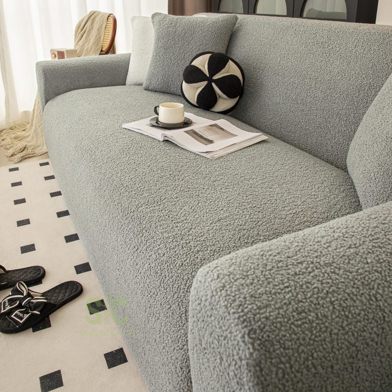 Op de afbeelding: Een lichtgrijze bankhoes met een getextureerd, fleece-achtig oppervlak. De hoes is over een bank geplaatst en bevat een bijpassend vierkant grijs sierkussen en een rond, zwart-wit patroon decoratief kussen op de zitkussens.