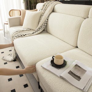 May include: A cream-colored, textured sherpa sofa cover draped over a couch. The cover features a soft, fleece-like material with a high-pile finish. A matching cream throw blanket with a black and white geometric pattern is layered on top.