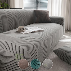 Könnte beinhalten: Ein grauer Sofabezug mit einem strukturierten schwarzen Fischgrätenmuster. Der Bezug ist über ein Sofa mit passendem Kissen gezogen. Ein offenes Buch und eine Keramiktasse liegen auf der Sitzfläche. Drei kreisförmige Farbmuster zeigen Braun, Grün und Grau.