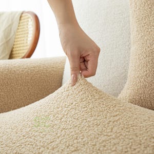 Op de afbeelding: Een beige sherpa stoffen bankhoes met een getextureerd, krullend oppervlak. Het materiaal is dik en zacht en heeft een consistente lichtbruine kleur. De stof wordt licht uitgerekt getoond om de flexibele, nauwsluitende aard te demonstreren.