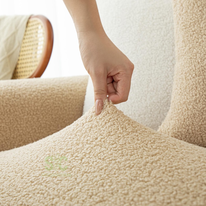 Op de afbeelding: Een beige sherpa stoffen bankhoes met een getextureerd, krullend oppervlak. Het materiaal is dik en zacht en heeft een consistente lichtbruine kleur. De stof wordt licht uitgerekt getoond om de flexibele, nauwsluitende aard te demonstreren.