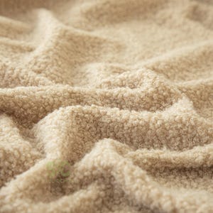 Op de afbeelding: Een beige stof met een dik, krullend en getextureerd oppervlak. Het materiaal vertoont diepe plooien en rimpelingen, wat zorgt voor een zachte en dichte uitstraling. Een klein lichtgroen logo met de tekst STUDIOCHUN is linksonder zichtbaar.